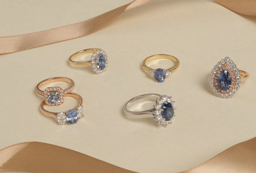 A collection of sapphire and diamond rings scattered across a beige background with a gold ribbon intertwined.