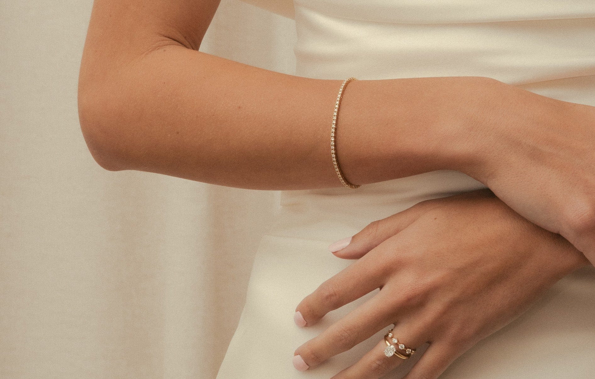 Bride in white gown with hands folded across her front, wearing a diamond set yellow gold tennis bracelet, oval cut engagement ring and diamond wedding ring.